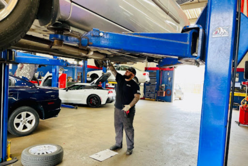 spring vehicle maintenance package at ABS Unlimited Auto Repair in Fairfax, VA. Image of mechanic standing under a silver car raised in the air on a rack doing an inspection of the front braking system with other cars around him in the shop bay.