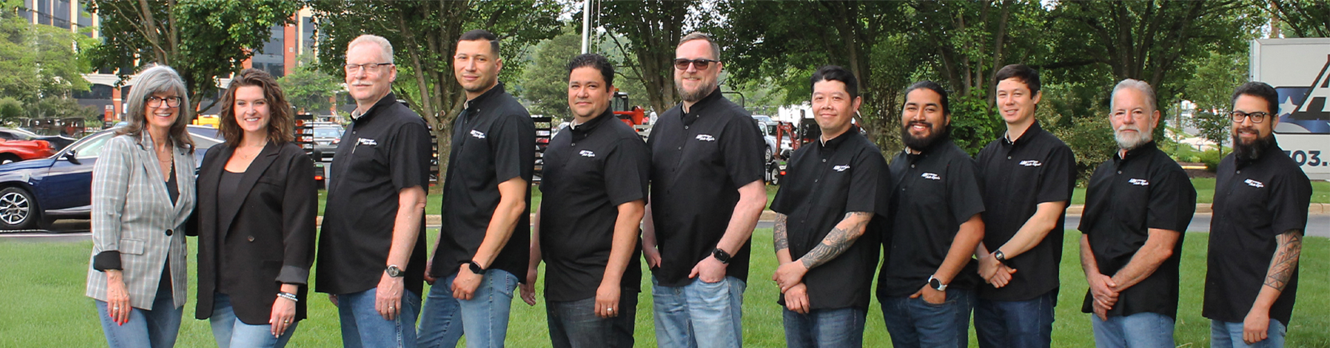 A group of eleven people standing outdoors in a line, most wearing matching black shirts with ABS Unlimited Auto Repair logo, in a park-like setting.